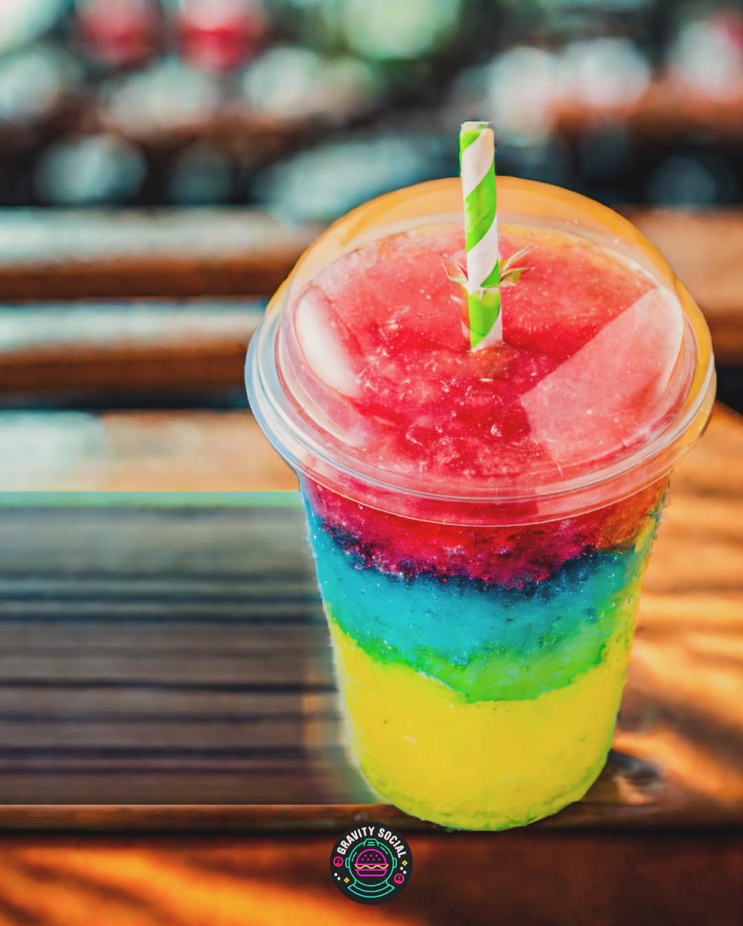Colorful layered slushie in a clear cup with a green-striped straw, placed on a wooden table.