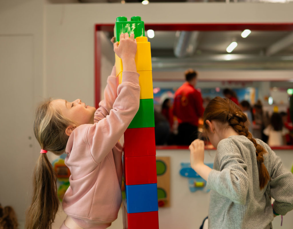 Two children build a tall structure with large colorful blocks in a playroom.
