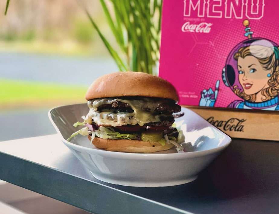 Cheeseburger on a white plate with lettuce and tomato, set against a colorful retro-themed menu backdrop.