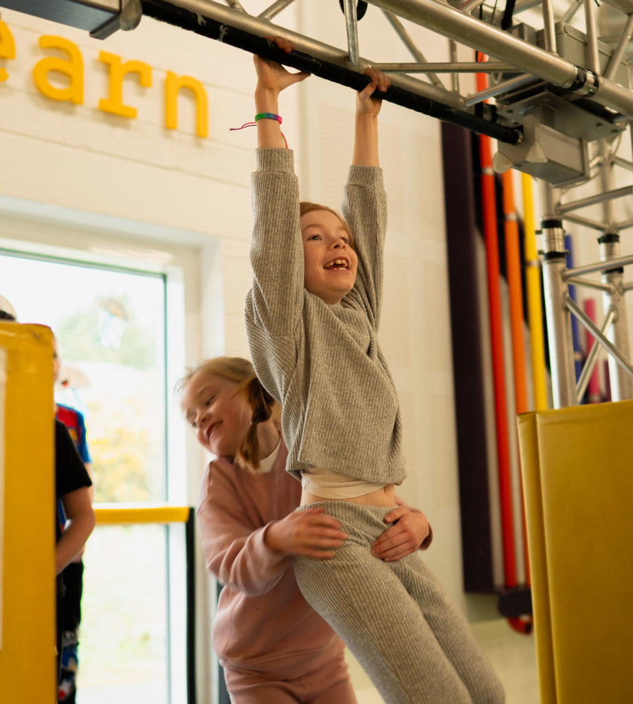 Child smiling while playing on indoor climbing structure, supported by a friend. Playful and energetic activity.