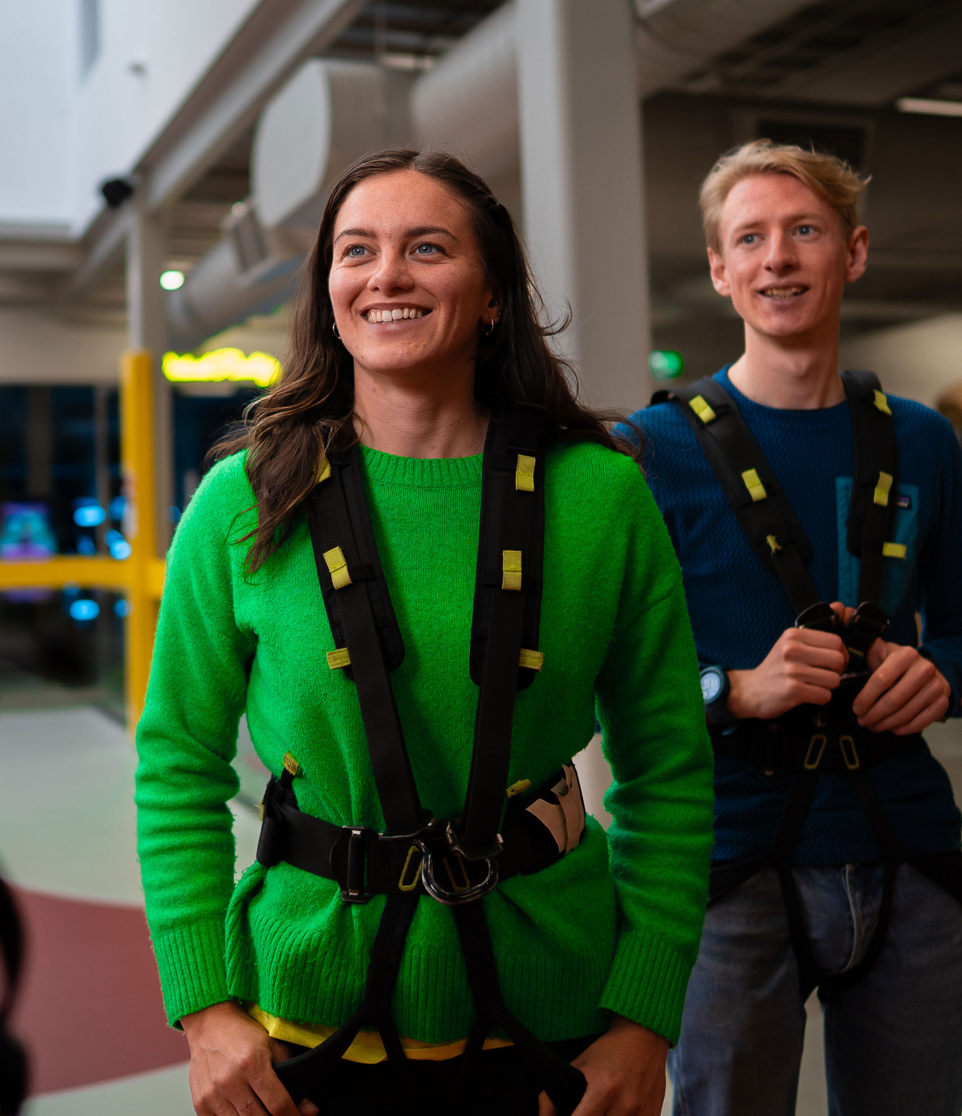 Two people in harnesses smiling, getting ready for an indoor adventure or climbing activity.