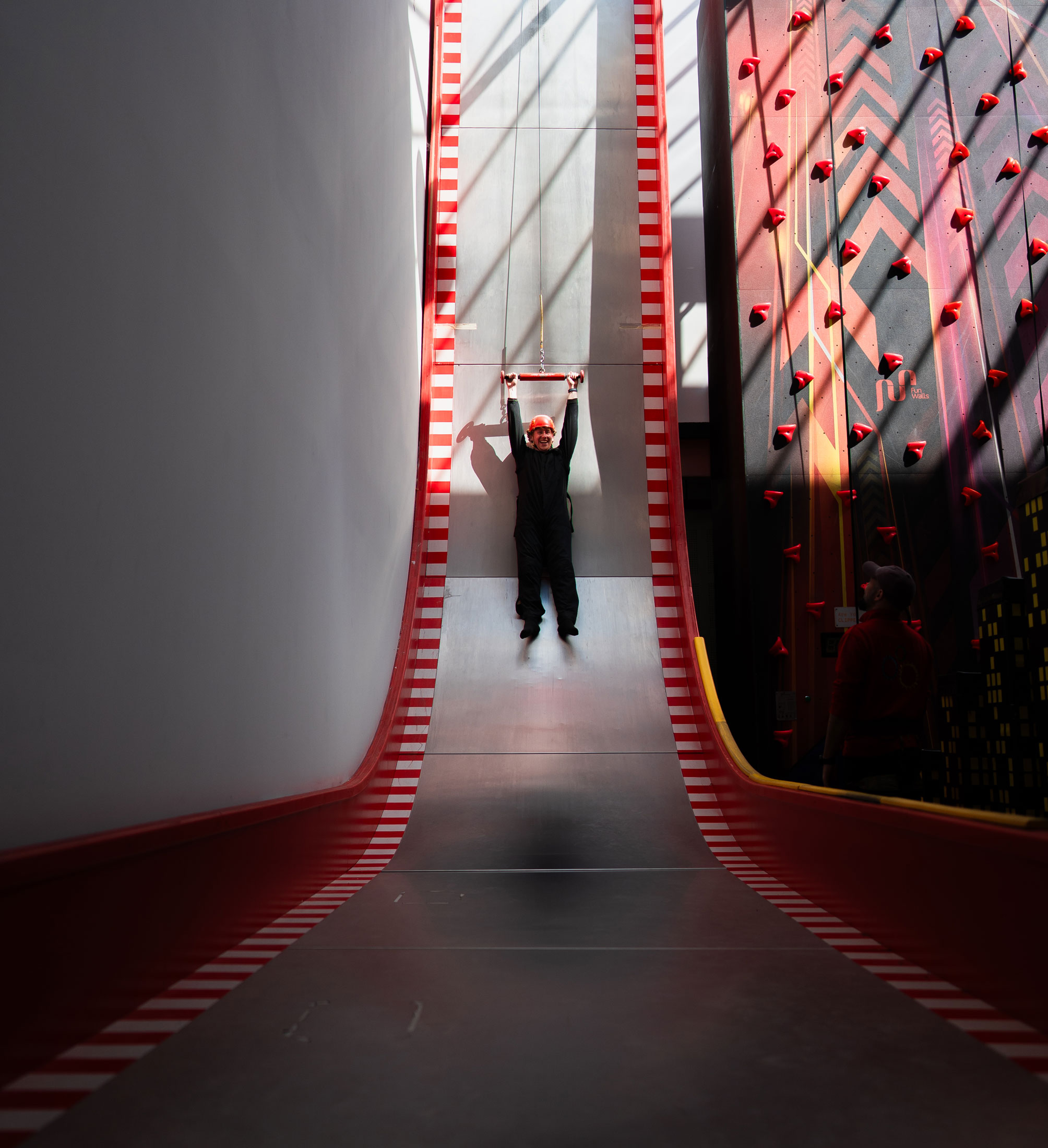 Person on vertical drop slide in adventure park, wearing safety gear, with climbing wall in background.